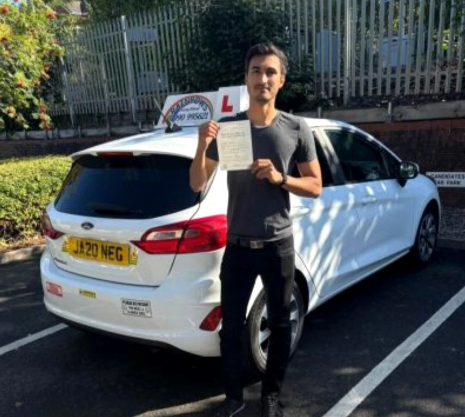 A proud Rainbows Driving School pupil holding a test pass certificate beside the white learner car